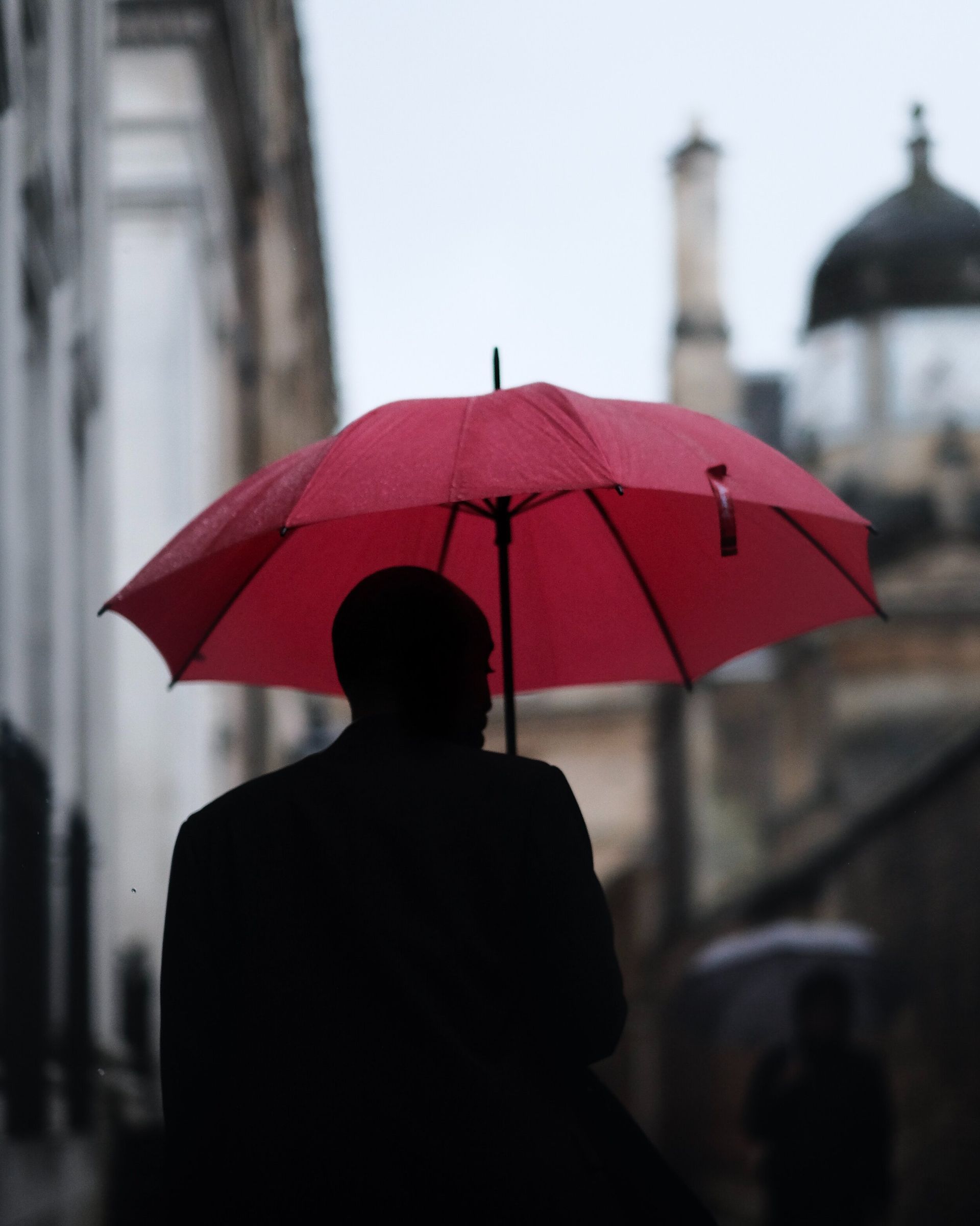 A person in a dark coat walking under a red umbrella on a rainy street