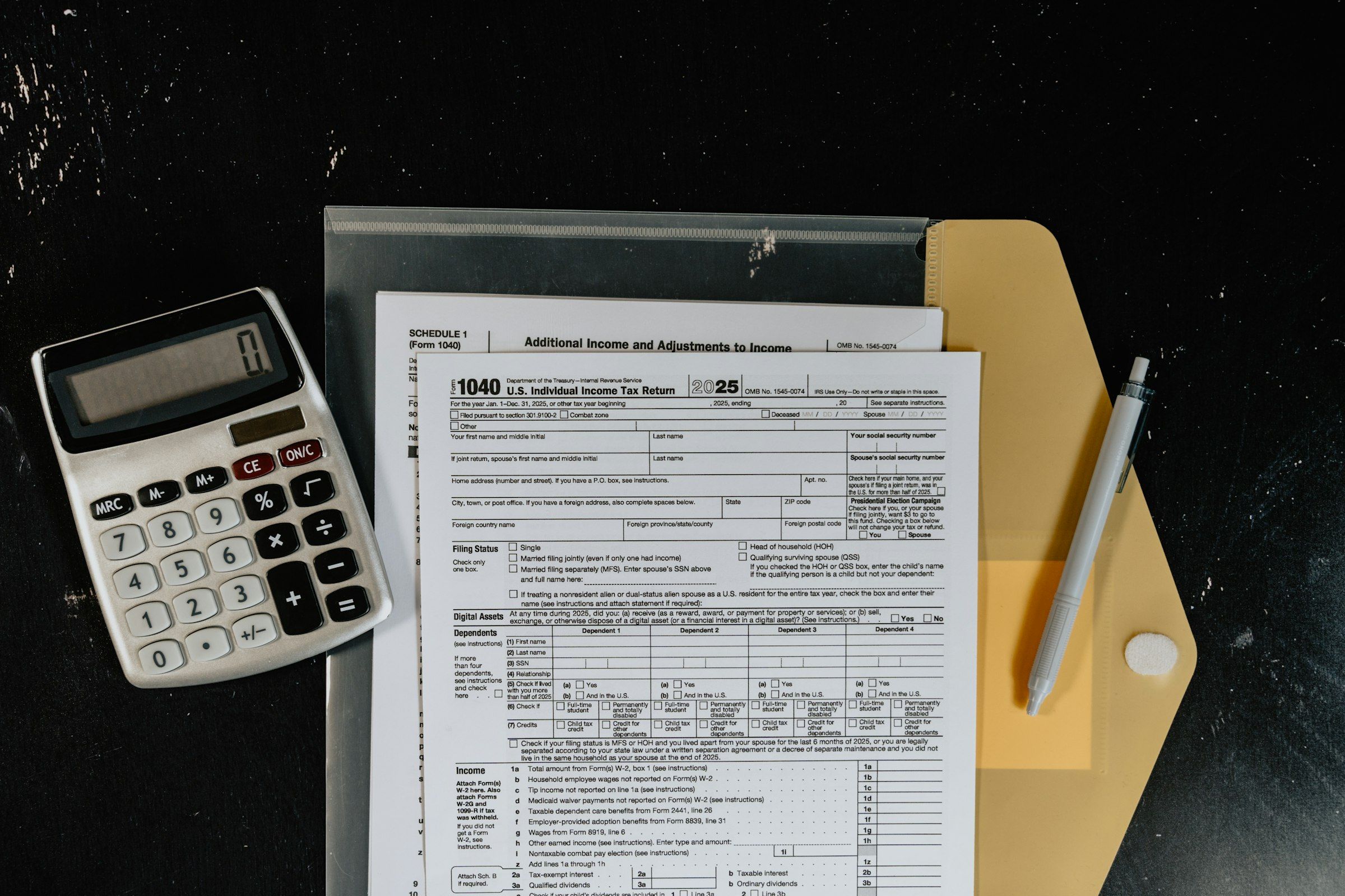 Form 1040 and Schedule 1 tax forms with a calculator, a pen, and a folder on a dark desk